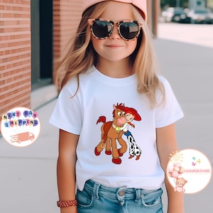 May include: White t-shirt featuring a cartoon graphic of a horse and a cowgirl hugging. The horse is brown with a saddle, and the cowgirl is wearing a red hat and blue jeans. The shirt is worn by a young person.
