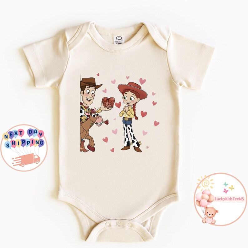 May include: Cream-colored baby onesie featuring a cartoon illustration of Woody and Jessie from Toy Story. Woody, on a horse, offers a heart-shaped box to Jessie, surrounded by pink hearts. The onesie has short sleeves and a round neckline.