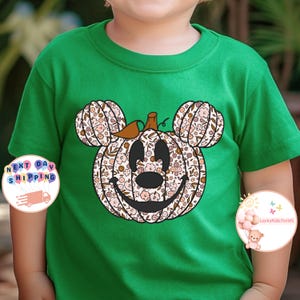 May include: A green t-shirt with a floral pumpkin Mickey Mouse design. The pumpkin is brown with a green stem and has a smiling face. The flowers are pink, white, and brown.