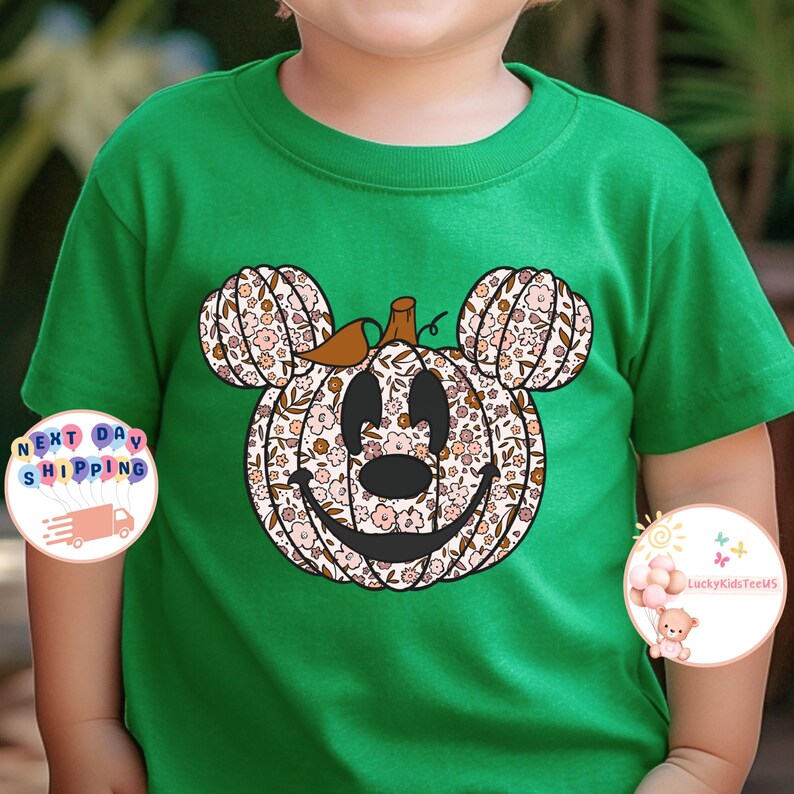 May include: A green t-shirt with a floral pumpkin Mickey Mouse design. The pumpkin is brown with a green stem and has a smiling face. The flowers are pink, white, and brown.