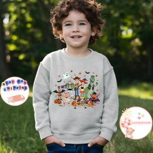 Puede incluir: Una sudadera gris claro con un gr&aacute;fico colorido de personajes de dibujos animados, incluyendo un fantasma, un vaquero, un guardabosques espacial y un dinosaurio. El dise&ntilde;o incluye murci&eacute;lagos, calabazas y hojas de oto&ntilde;o. La sudadera la lleva un ni&ntilde;o peque&ntilde;o.