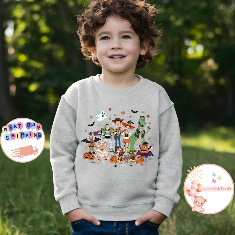 Puede incluir: Una sudadera gris claro con un gr&aacute;fico colorido de personajes de dibujos animados, incluyendo un fantasma, un vaquero, un guardabosques espacial y un dinosaurio. El dise&ntilde;o incluye murci&eacute;lagos, calabazas y hojas de oto&ntilde;o. La sudadera la lleva un ni&ntilde;o peque&ntilde;o.