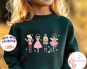 Könnte beinhalten: Dunkelgrünes Sweatshirt mit einem Nussknacker-Themen-Design. Die Grafik zeigt eine Maus, eine Ballerina, einen Nussknacker und eine Fee in festlicher Kleidung. Das Design ist mit Sternen und einem kleinen Weihnachtsbaum verziert.