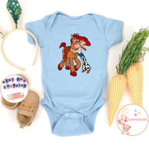 May include: A light blue baby onesie featuring a cartoon image of Jessie and Bullseye from Toy Story. The illustration shows Jessie hugging Bullseye. The onesie has short sleeves and a snap closure at the bottom. The onesie is surrounded by Easter-themed decorations.