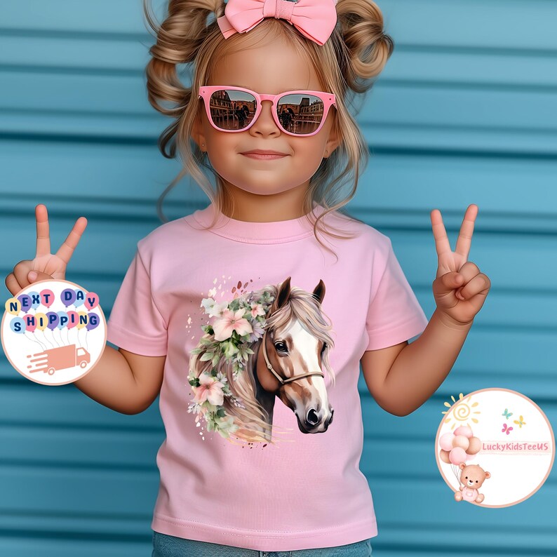 May include: A pink t-shirt featuring a watercolor illustration of a horse's head adorned with flowers. The image is on a young child, who is wearing pink sunglasses and a pink bow. The background is a blue wall.