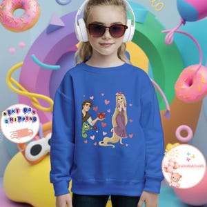 May include: A blue sweatshirt featuring a cartoon illustration of Rapunzel and Flynn Rider with hearts. The sweatshirt is worn by a person wearing sunglasses and headphones. Pink and yellow donuts are in the background.