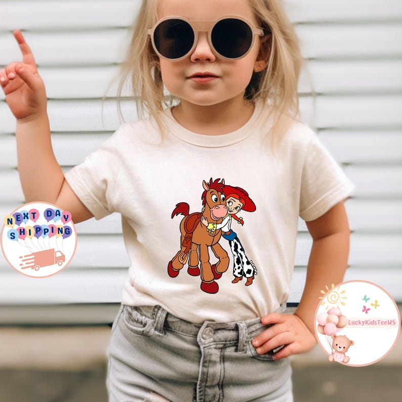 May include: A beige t-shirt featuring a cartoon graphic of a horse and a cowgirl hugging. The horse is brown with a saddle, and the cowgirl has red hair and a blue and white outfit. The shirt is for children.