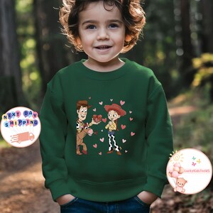 May include: A dark green sweatshirt featuring a cartoon illustration of two characters, a cowboy and a cowgirl, with hearts. The cowboy is on a horse and offering a gift to the cowgirl. The sweatshirt is worn by a child.