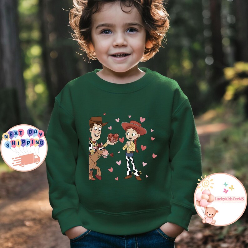 May include: A dark green sweatshirt featuring a cartoon illustration of two characters, a cowboy and a cowgirl, with hearts. The cowboy is on a horse and offering a gift to the cowgirl. The sweatshirt is worn by a child.