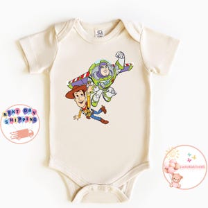 May include: Cream-colored baby bodysuit featuring a colorful graphic of Woody and Buzz Lightyear from Toy Story. Woody is wearing a cowboy hat and Buzz has his wings extended. The bodysuit has short sleeves and a round neckline.