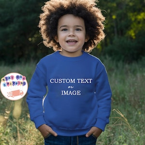 May include: A young child wearing a royal blue sweatshirt with the words "CUSTOM TEXT or IMAGE" in white. The child is smiling, with hands in pockets. The background is a blurred green and brown outdoor scene.
