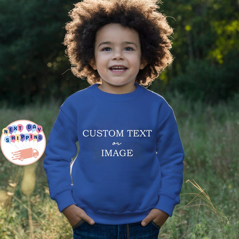 May include: A young child wearing a royal blue sweatshirt with the words "CUSTOM TEXT or IMAGE" in white. The child is smiling, with hands in pockets. The background is a blurred green and brown outdoor scene.
