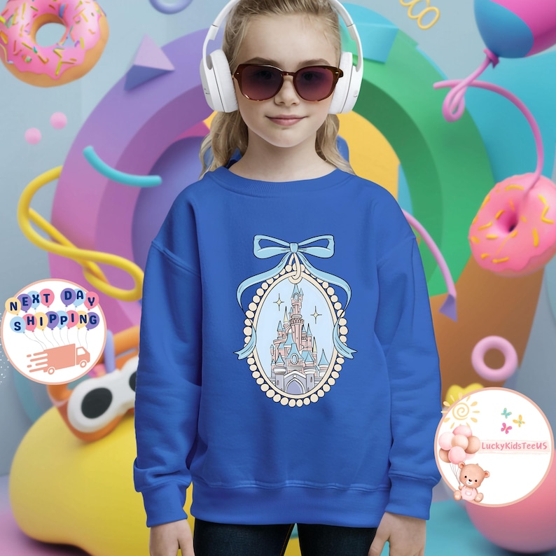 Puede incluir: Una sudadera azul real con una ilustraci&oacute;n azul claro y rosa de un castillo con un dise&ntilde;o de perlas y cinta. El castillo est&aacute; rodeado por un borde de perlas.