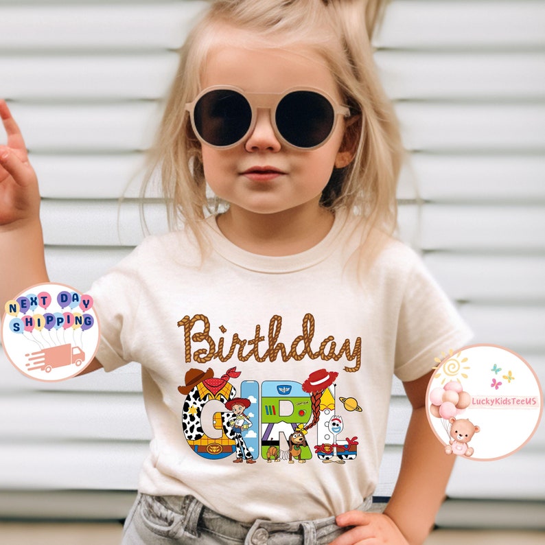 Op de afbeelding: Een beige T-shirt met een bruin touwontwerp met de tekst "Birthday Girl". De letters zijn gemaakt van afbeeldingen van personages uit de Toy Story-filmfranchise.