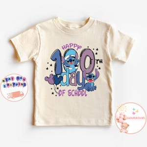 May include: A light beige t-shirt with a blue and purple graphic of Stitch characters celebrating the 100th day of school. The text "Happy 100th day of school" is printed on the shirt.