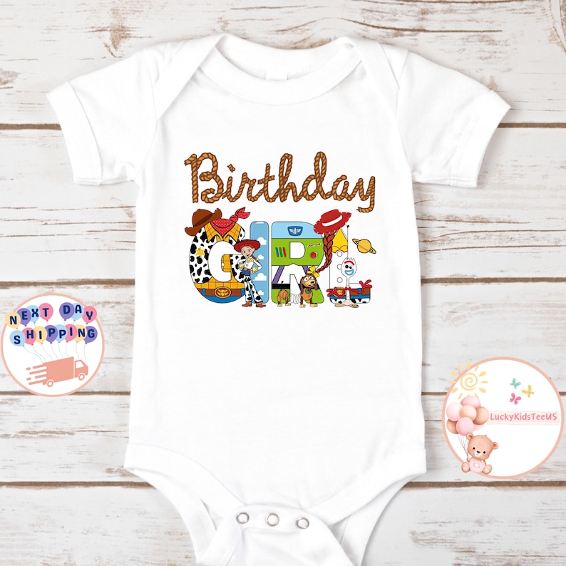 Op de afbeelding: Witte romper met een bruin touwontwerp rond het woord "Birthday" en het woord "Girl" gespeld met personages uit de Toy Story-film. De personages zijn Woody, Jessie, Buzz Lightyear en Bullseye.