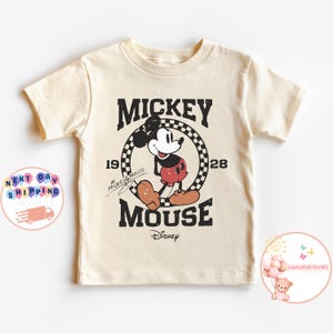 K&ouml;nnte beinhalten: Ein cremefarbenes T-Shirt mit einem Vintage-Mickey-Mouse-Motiv. Das Motiv zeigt Mickey Mouse in Schwarz und Rot, der in einem karierten Kreis steht, mit dem Text "Mickey Mouse" und "1928" dar&uuml;ber und "Mouse" darunter. Der Text "Disney" befindet sich unter dem Wort "Mouse".