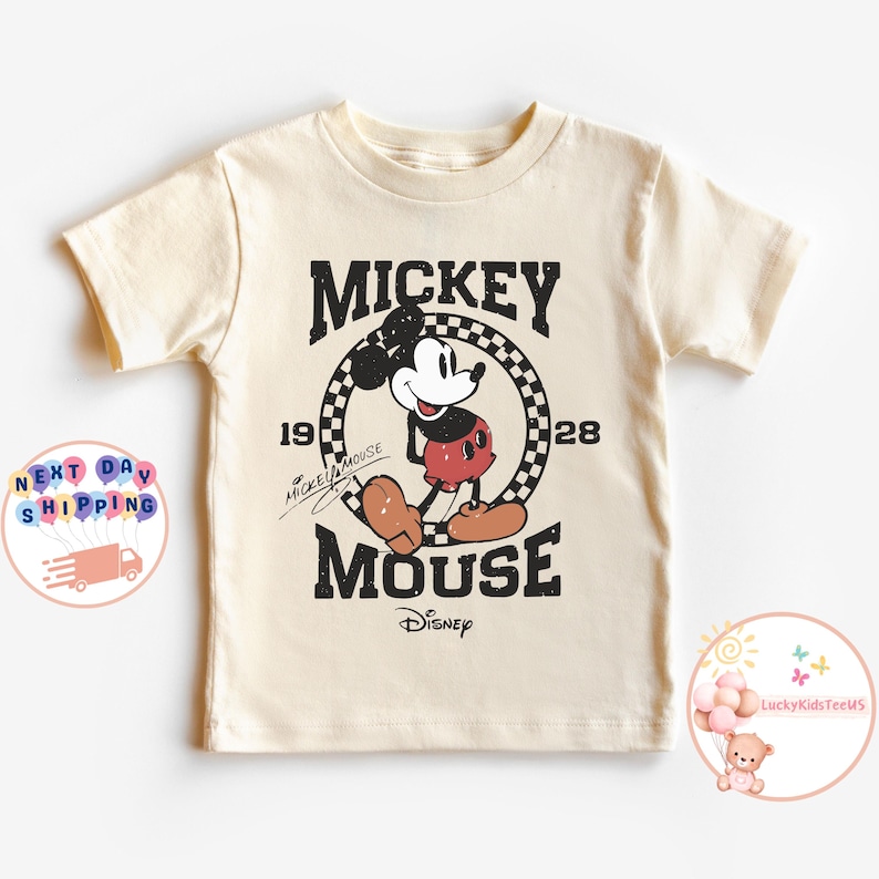 K&ouml;nnte beinhalten: Ein cremefarbenes T-Shirt mit einem Vintage-Mickey-Mouse-Motiv. Das Motiv zeigt Mickey Mouse in Schwarz und Rot, der in einem karierten Kreis steht, mit dem Text "Mickey Mouse" und "1928" dar&uuml;ber und "Mouse" darunter. Der Text "Disney" befindet sich unter dem Wort "Mouse".