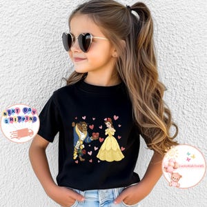 May include: Black t-shirt featuring a cartoon illustration of Belle and the Beast with heart accents. Belle is wearing a yellow dress, and the Beast is in a blue coat. The shirt is worn by a young person.