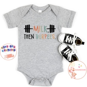 May include: A gray baby onesie with the text "MILK, THEN BURPEES" in black, peach, green, and blue. The design includes a barbell graphic. Two small black and white sneakers are next to the onesie.