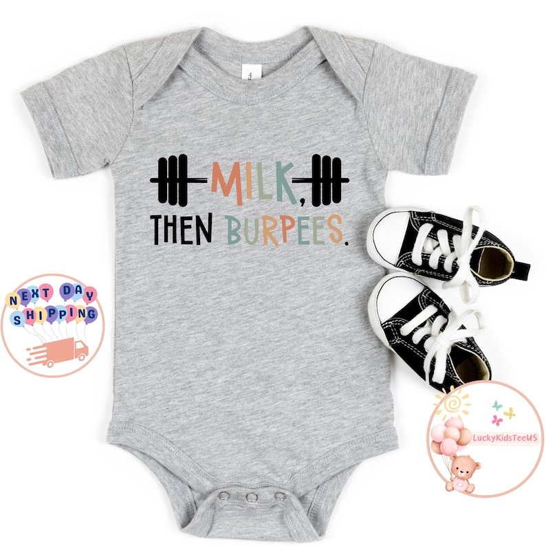 May include: A gray baby onesie with the text "MILK, THEN BURPEES" in black, peach, green, and blue. The design includes a barbell graphic. Two small black and white sneakers are next to the onesie.
