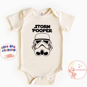 May include: A white baby onesie with a black graphic of a Star Wars Stormtrooper helmet and the text "Storm Pooper".