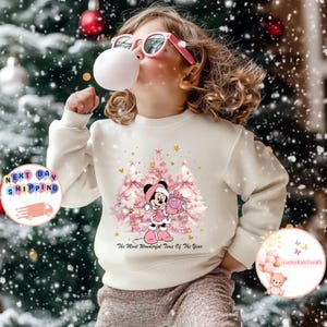 May include: A cream-colored sweatshirt featuring a graphic of Minnie Mouse in a pink outfit holding a gift box in front of pink Christmas trees. The text "The Most Wonderful Time Of The Year" is below the graphic. A child is wearing the sweatshirt.