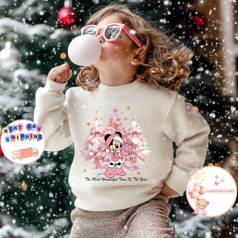 May include: A cream-colored sweatshirt featuring a graphic of Minnie Mouse in a pink outfit holding a gift box in front of pink Christmas trees. The text "The Most Wonderful Time Of The Year" is below the graphic. A child is wearing the sweatshirt.