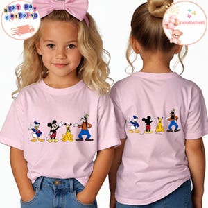 Op de afbeelding: Twee jonge meisjes dragen lichtroze t-shirts met een cartoonafbeelding van Disney-personages. De voorkant van de shirts toont Donald Duck, Mickey Mouse, Pluto en Goofy. De achterkant van de shirts toont dezelfde personages. De meisjes dragen een blauwe jeans.