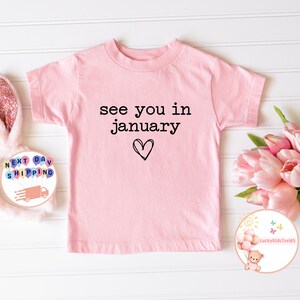 May include: Pink short-sleeved t-shirt with the text "see you in january" and a heart drawn underneath.