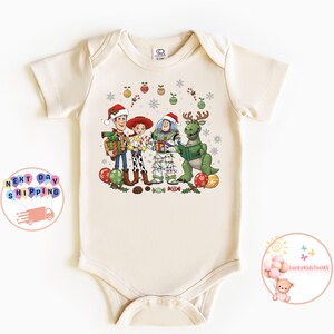 May include: A cream-coloured baby bodysuit with a festive graphic of Toy Story characters wearing Santa hats and surrounded by Christmas decorations. The design includes Woody, Jessie, Buzz Lightyear, and Rex, ideal for the holiday season.