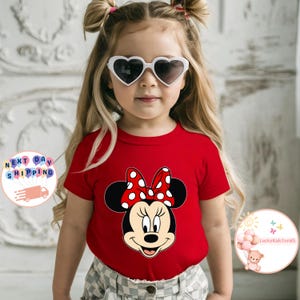 May include: A red t-shirt with a Minnie Mouse design, including a polka-dot bow. The shirt is worn by a young child wearing heart-shaped sunglasses. The child has blonde hair styled in pigtails.