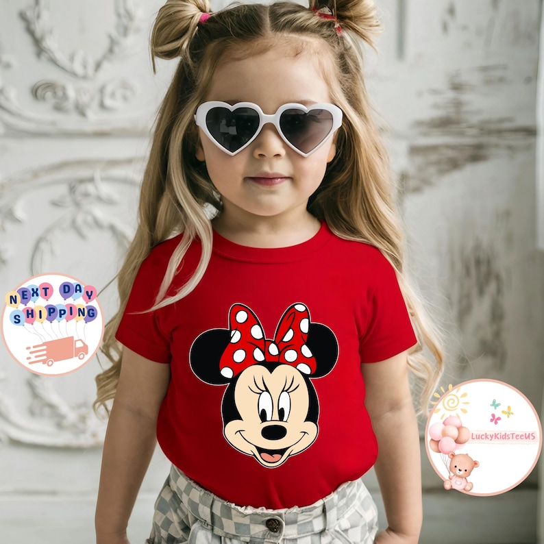 May include: A red t-shirt with a Minnie Mouse design, including a polka-dot bow. The shirt is worn by a young child wearing heart-shaped sunglasses. The child has blonde hair styled in pigtails.