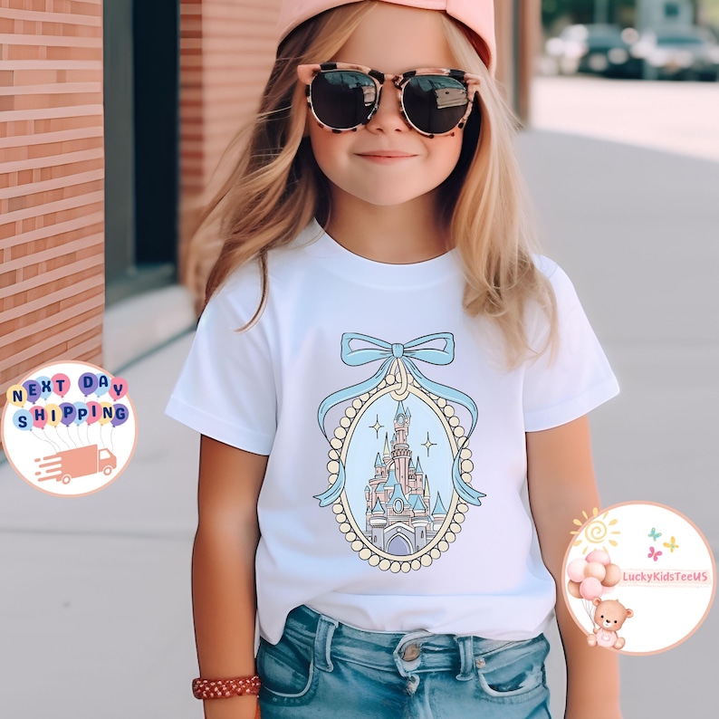 Puede incluir: Una camiseta blanca con una ilustraci&oacute;n azul y rosa de un castillo en un marco de perlas y cinta. El castillo est&aacute; rodeado de estrellas.