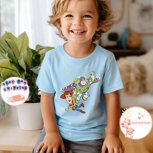 May include: A light blue short-sleeve t-shirt featuring a graphic of Buzz Lightyear and Woody from Toy Story. The cartoon characters are in mid-air. The shirt is worn by a smiling child.