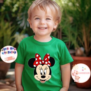 May include: A green t-shirt with a Minnie Mouse design, including a red bow with white spots. The shirt is worn by a young child. The image also includes the text "Next Day Shipping" and "LuckyKidsTeeUS".