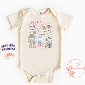 May include: A cream-coloured baby onesie with Star Wars illustrations. The design includes a Stormtrooper, R2-D2, and the Mandalorian helmet, along with the phrase "May the Force Be With You".