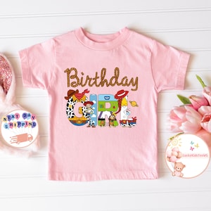 Op de afbeelding: Roze T-shirt met korte mouwen met de tekst "Birthday Girl" in bruine touwletters. De letters zijn versierd met personages uit de Toy Story-filmfranchise.