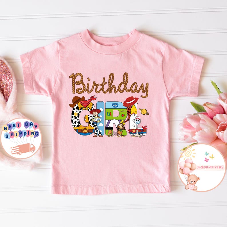 Op de afbeelding: Roze T-shirt met korte mouwen met de tekst "Birthday Girl" in bruine touwletters. De letters zijn versierd met personages uit de Toy Story-filmfranchise.
