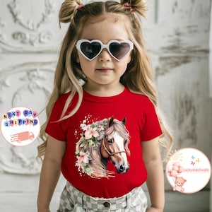 May include: A red t-shirt featuring a watercolor illustration of a horse's head adorned with flowers. The horse has a brown and white coat and a bridle. The shirt is worn by a young child wearing heart-shaped sunglasses.