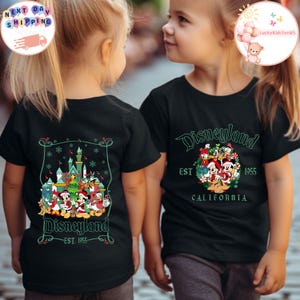 May include: Two children wearing black t-shirts with a Christmas-themed Disneyland graphic. The front of the shirt says "Disneyland EST. 1955 CALIFORNIA". The back of the shirt features a Christmas tree with Mickey Mouse, Minnie Mouse, Donald Duck, Goofy, Pluto, and other Disney characters.