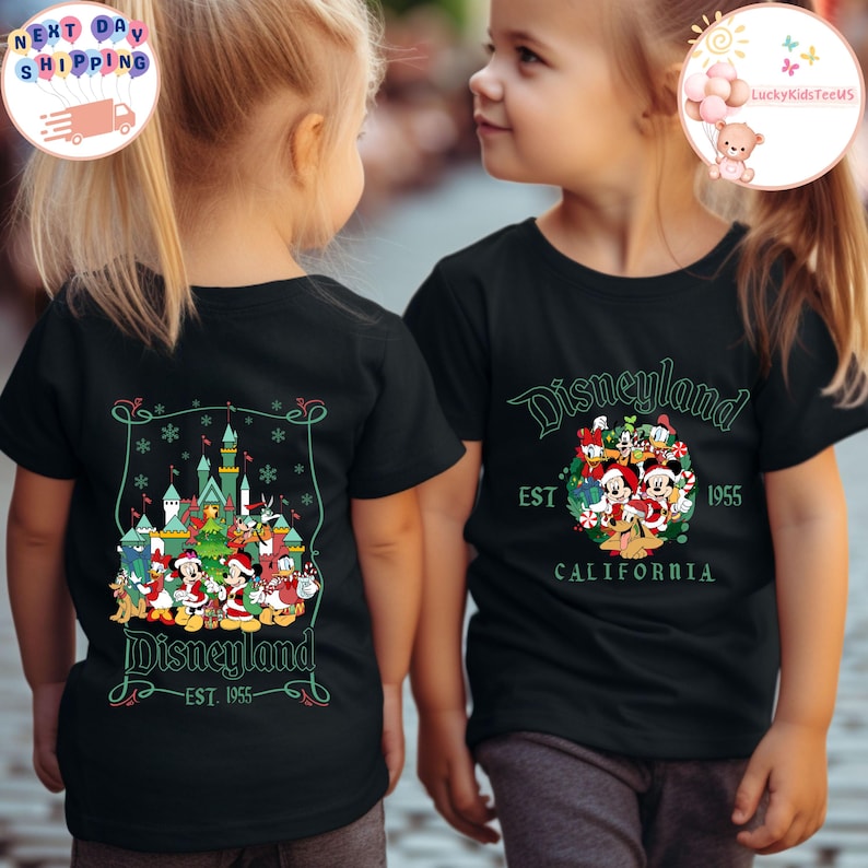 May include: Two children wearing black t-shirts with a Christmas-themed Disneyland graphic. The front of the shirt says "Disneyland EST. 1955 CALIFORNIA". The back of the shirt features a Christmas tree with Mickey Mouse, Minnie Mouse, Donald Duck, Goofy, Pluto, and other Disney characters.