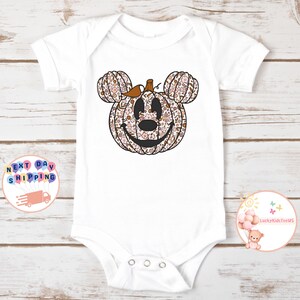 May include: White baby onesie with a floral pumpkin Mickey Mouse design. The pumpkin is brown and orange with pink and white flowers.