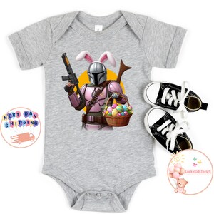 May include: A gray baby onesie with a graphic of a Mandalorian character wearing bunny ears and holding a basket of Easter eggs. The character is holding a blaster in one hand and a basket of Easter eggs in the other.