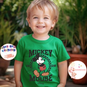 K&ouml;nnte beinhalten: Gr&uuml;nes T-Shirt mit kurzen &Auml;rmeln und einem schwarz-wei&szlig;en Motiv von Mickey Mouse. Das Motiv lautet "Mickey Mouse" und hat die Zahlen "1928" au&szlig;en herum. Das Shirt hat auch den Text "Disney" unten.