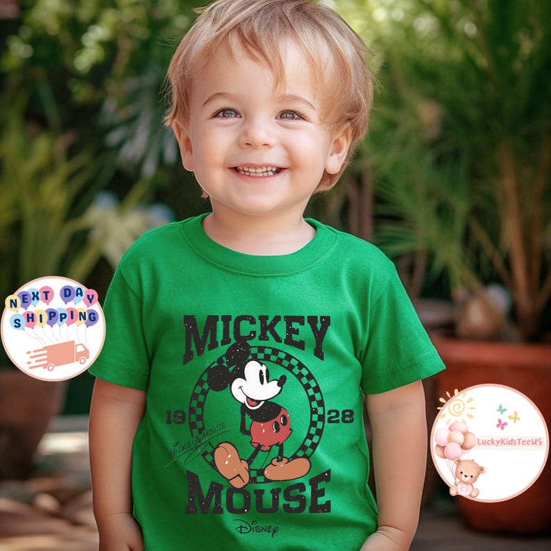 K&ouml;nnte beinhalten: Gr&uuml;nes T-Shirt mit kurzen &Auml;rmeln und einem schwarz-wei&szlig;en Motiv von Mickey Mouse. Das Motiv lautet "Mickey Mouse" und hat die Zahlen "1928" au&szlig;en herum. Das Shirt hat auch den Text "Disney" unten.