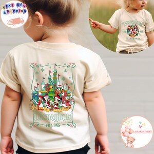 May include: A beige t-shirt with a colorful graphic of the Disneyland castle and characters. The text "Disneyland - EST. 1955" is printed below the graphic.