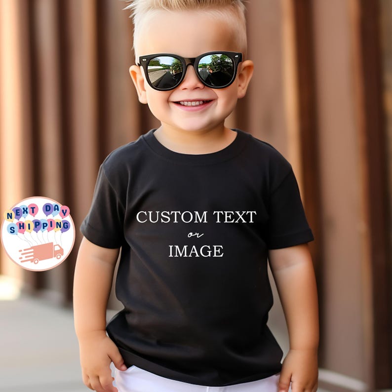 May include: A young child wearing a black t-shirt with the words "CUSTOM TEXT or IMAGE" in white. The child is also wearing black sunglasses and smiling. The background is a blurred brown.