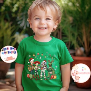 May include: A green t-shirt with a Christmas-themed Toy Story graphic. The design includes Woody, Jessie, Buzz Lightyear, and Rex in festive attire, surrounded by Christmas decorations. The child is smiling.