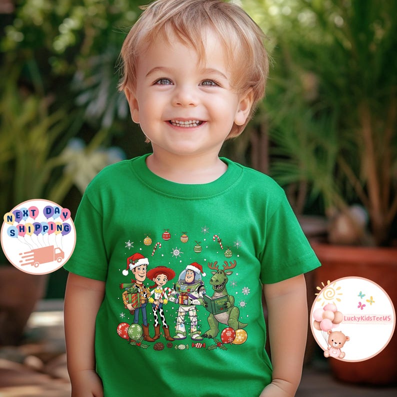 May include: A green t-shirt with a Christmas-themed Toy Story graphic. The design includes Woody, Jessie, Buzz Lightyear, and Rex in festive attire, surrounded by Christmas decorations. The child is smiling.